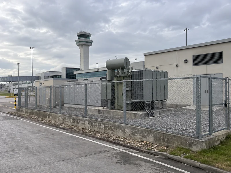 Fenced transformer utility yard with an airport terminal and control tower behind it