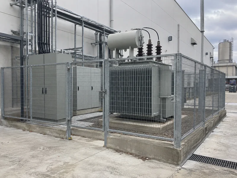 Outdoor transformer in a fenced utility yard beside an industrial factory building