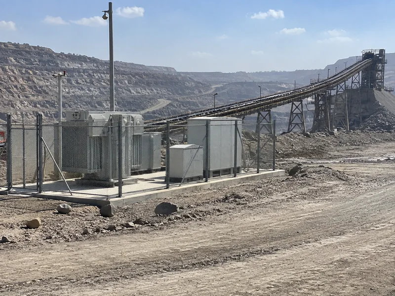 Fenced transformer and electrical cabinets beside an ore conveyor at a surface mine